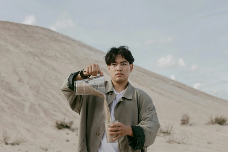 a man standing in the desert pouring something into a glass, an album cover, by Robbie Trevino, pexels contest winner, male ulzzang, iced latte, half asian, confident looking