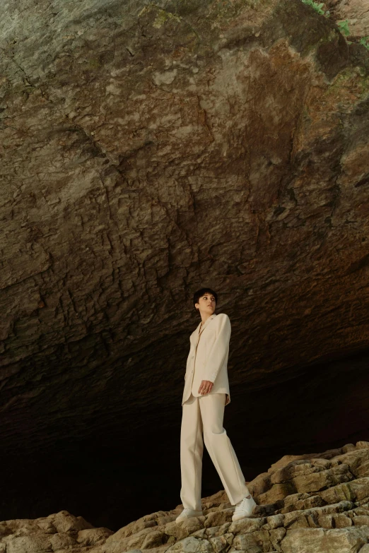 a woman standing in front of a large rock, inspired by Vanessa Beecroft, unsplash, renaissance, wearing white suit, androgynous male, wearing cave man clothes, official store photo