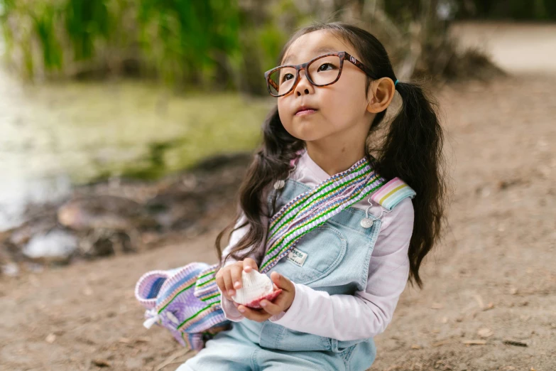 a little girl sitting on the ground next to a body of water, inspired by Hikari Shimoda, trending on pexels, square rimmed glasses, aboriginal australian hipster, having a picnic, closeup of an adorable