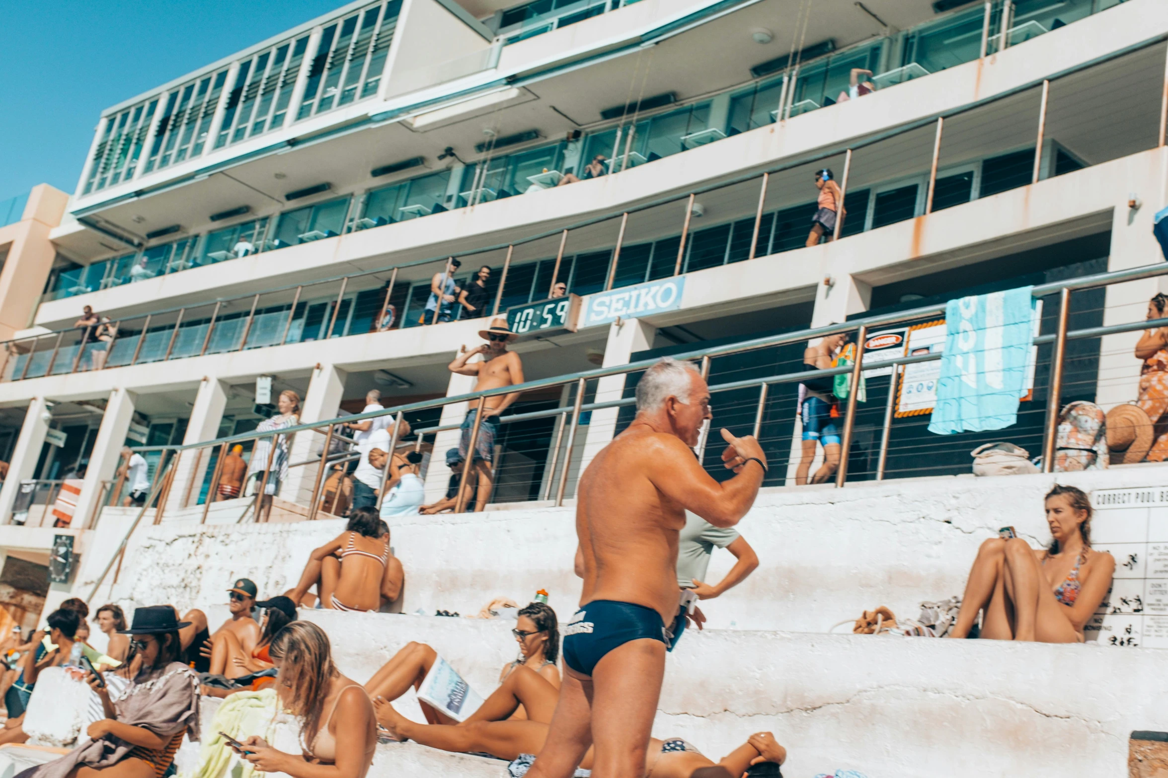 a man standing on top of a beach next to a crowd of people, pexels contest winner, figuration libre, sit on the edge of swimming pool, bondi beach in the background, art deco stadium, boutinela bikini