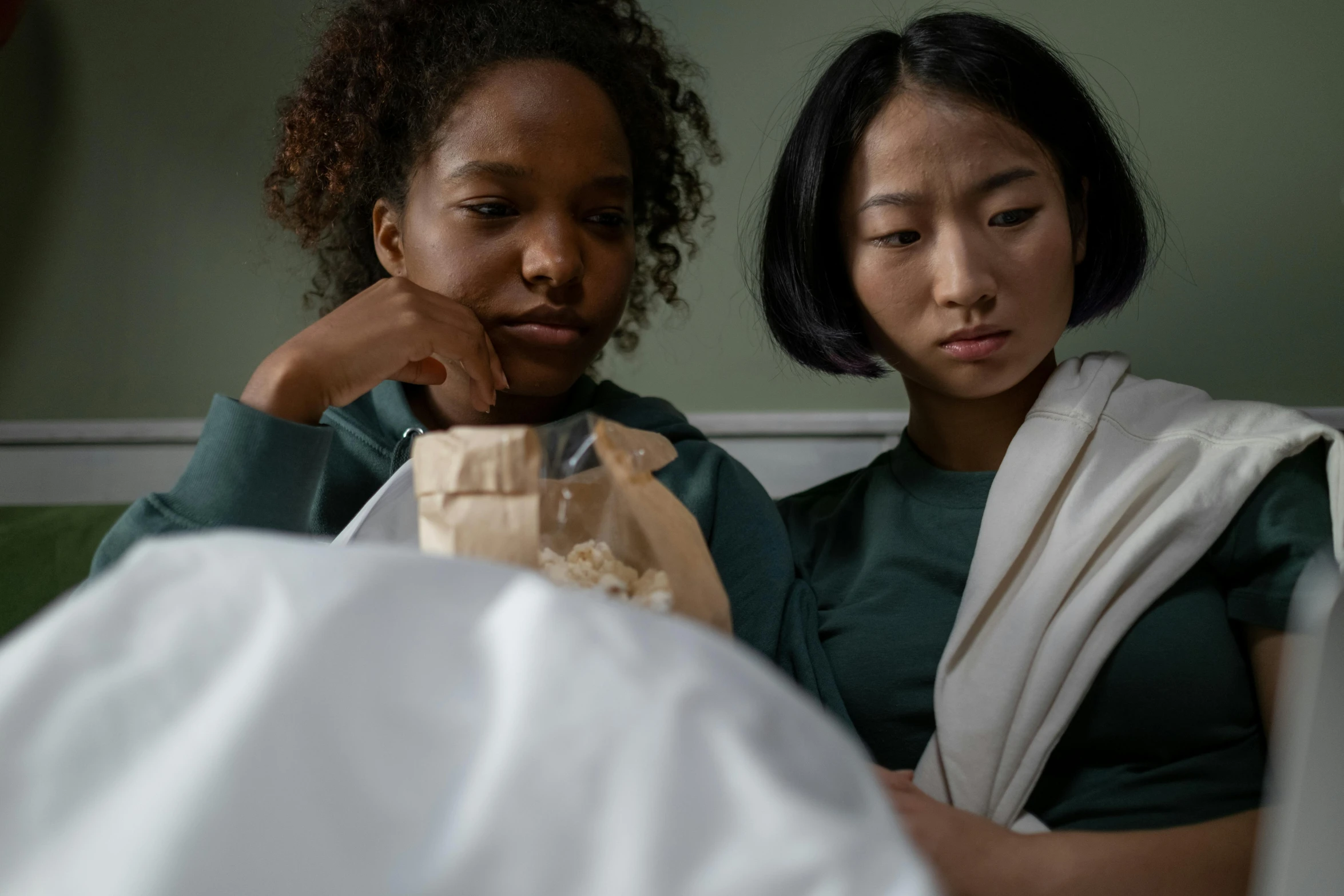 a couple of women sitting on top of a bed, inspired by Zhang Xiaogang, trending on pexels, at the hospital in patient gown, eating chips and watching tv, sorrow, mixed race