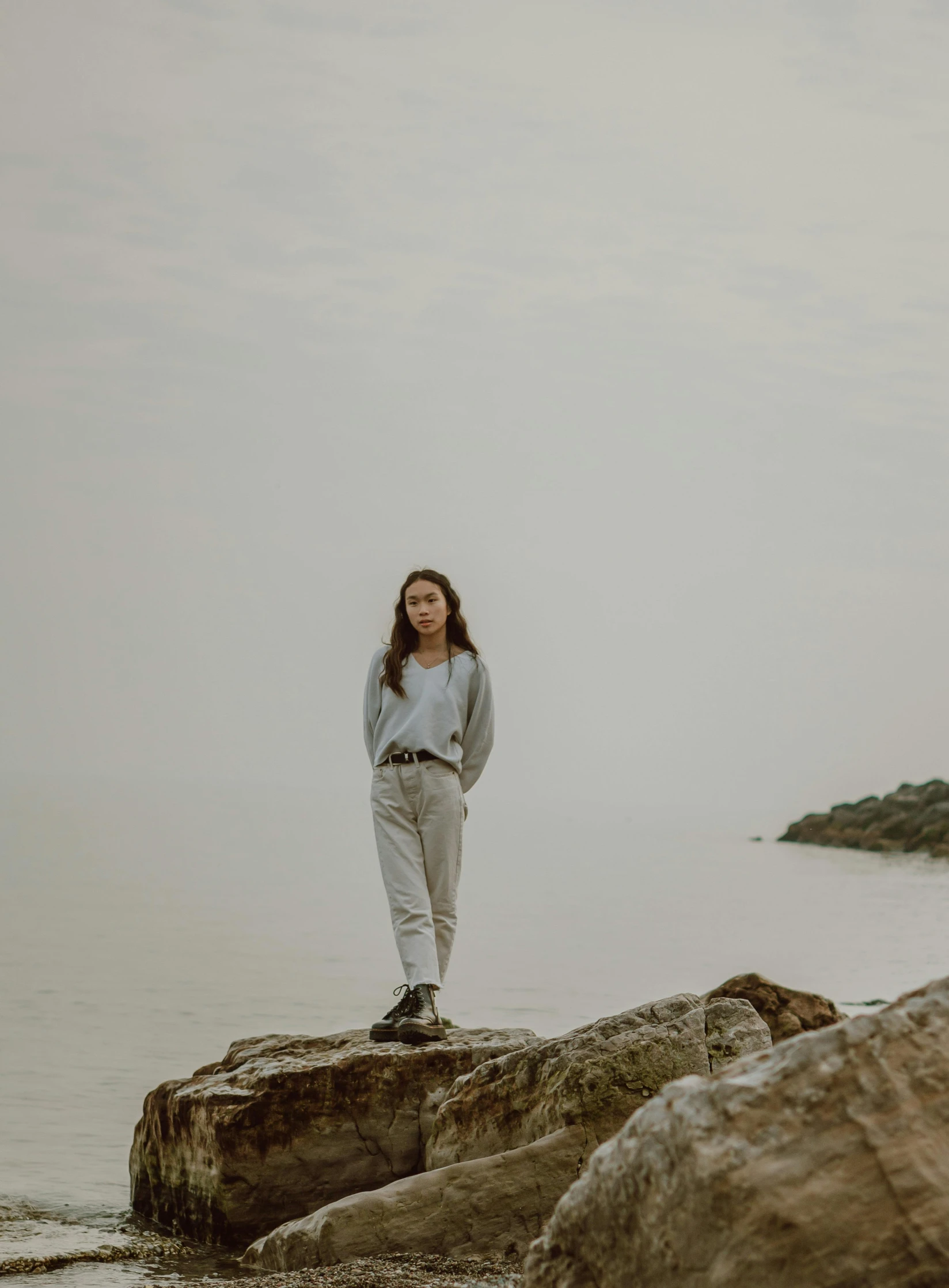 a woman standing on a rock in front of a body of water, profile image, gray canvas, wearing casual clothes, trending photo