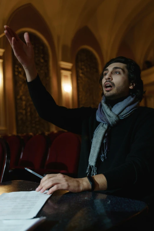 a man sitting at a table with papers in front of him, singing at a opera house, adam driver, amr elshamy, epk