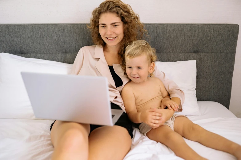 a woman and child sitting on a bed with a laptop, a cartoon, pexels, casually dressed, attractive photo, close - up photograph, gold