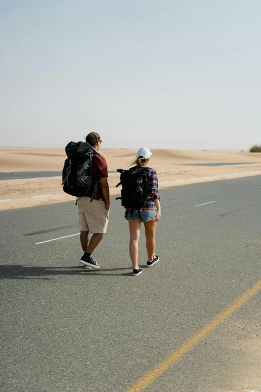 a couple of people walking down a road, in the desert beside the gulf, with a backpack, wearing shorts, paved