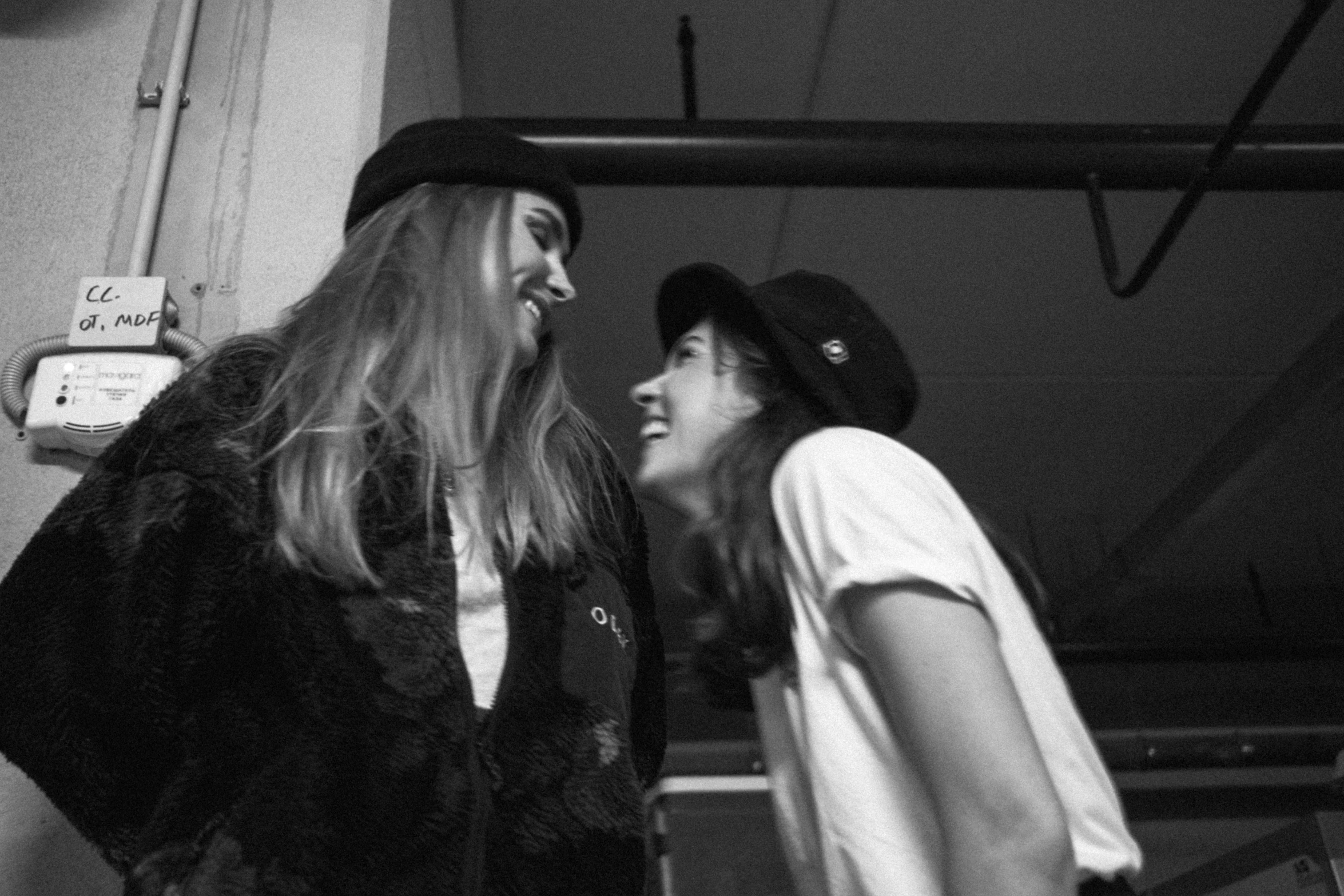 a couple of women standing next to each other, a black and white photo, tumblr, happening, smiling at each other, caps sideways, warpaint aesthetic, indoor picture