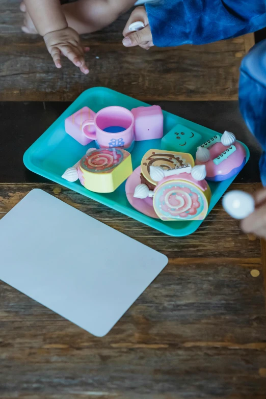 a small child sitting on top of a wooden table, a child's drawing, inspired by Gerard David, unsplash, marshmallows, teal and pink, cups and balls, square