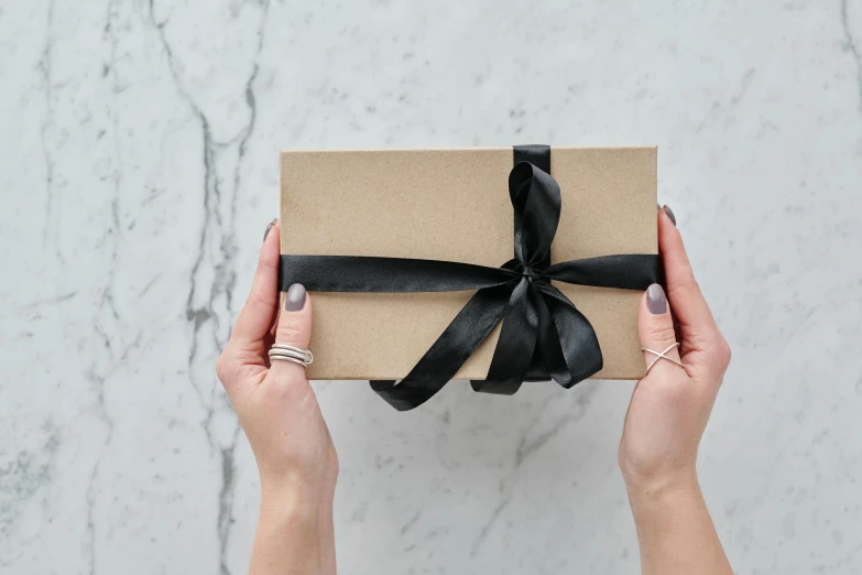 a person holding a brown box with a black ribbon, by Eden Box, pexels contest winner, private press, all marble, satin silver, thumbnail, angled shot