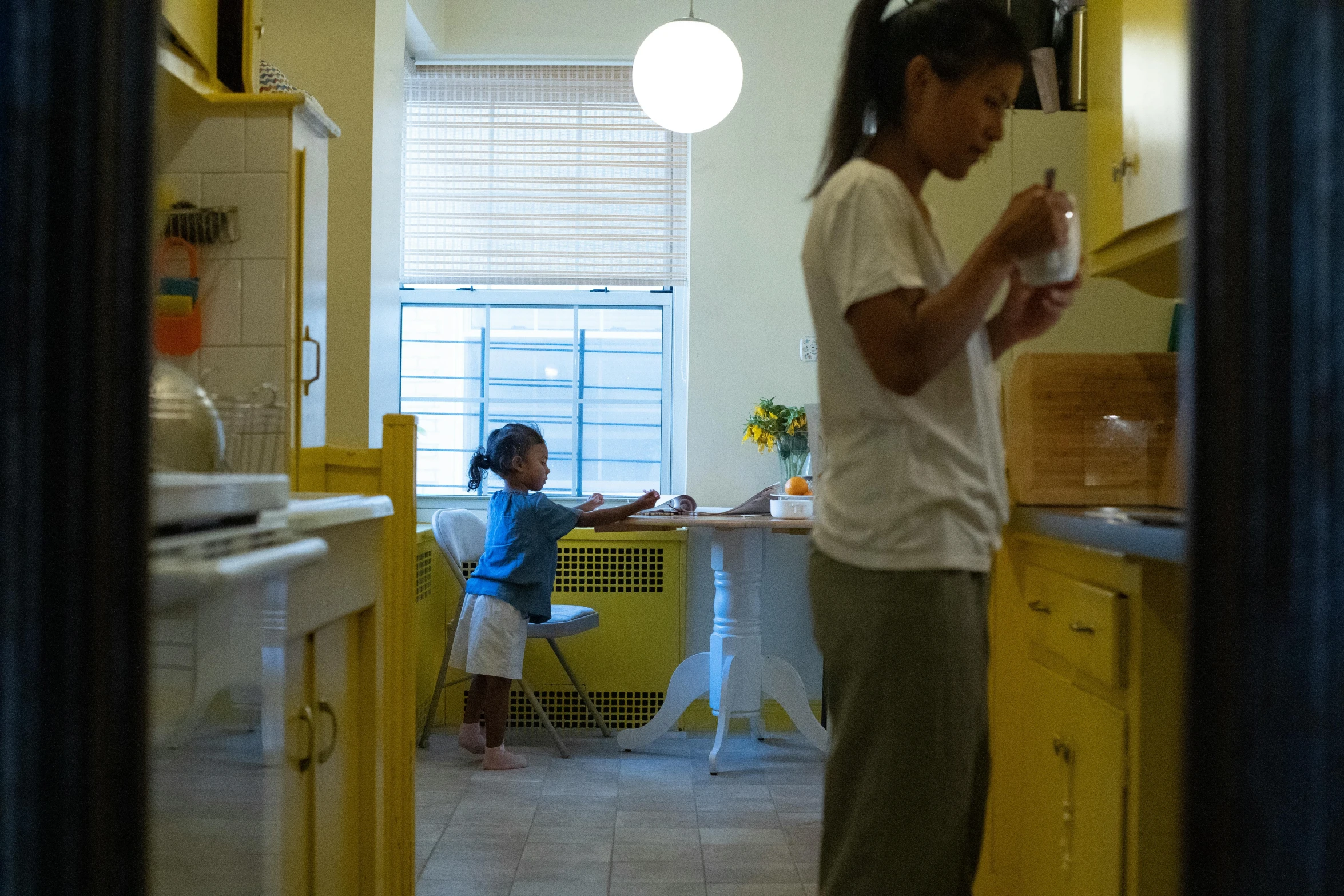 a woman standing in a kitchen next to a little girl, a picture, bright yellow color scheme, ignant, contaning tables and walls, distant photo