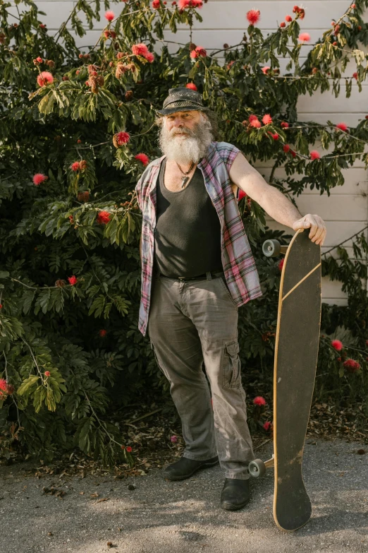 a man standing in front of a bush holding a skateboard, grey beard, profile image, hillbilly, noelle stevenson