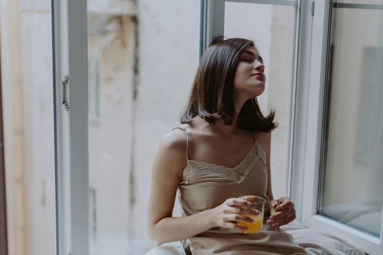 a woman sitting on a window sill holding a drink, by Emma Andijewska, trending on pexels, renaissance, background image, wearing a camisole, honey, clean face and body skin