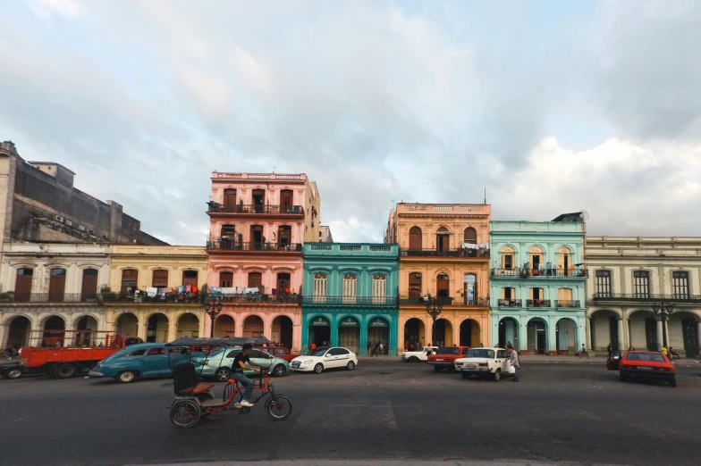 a group of cars driving down a street next to tall buildings, a colorized photo, pexels contest winner, hyperrealism, cuba, colorful houses, square, early evening