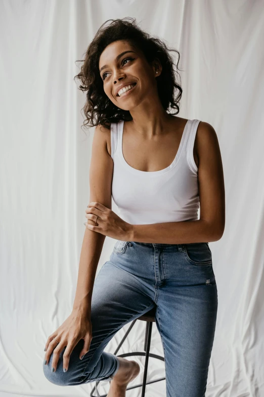 a woman sitting on a stool in front of a white backdrop, trending on pexels, outfit : jeans and white vest, wearing : tanktop, small wide smile, ashteroth