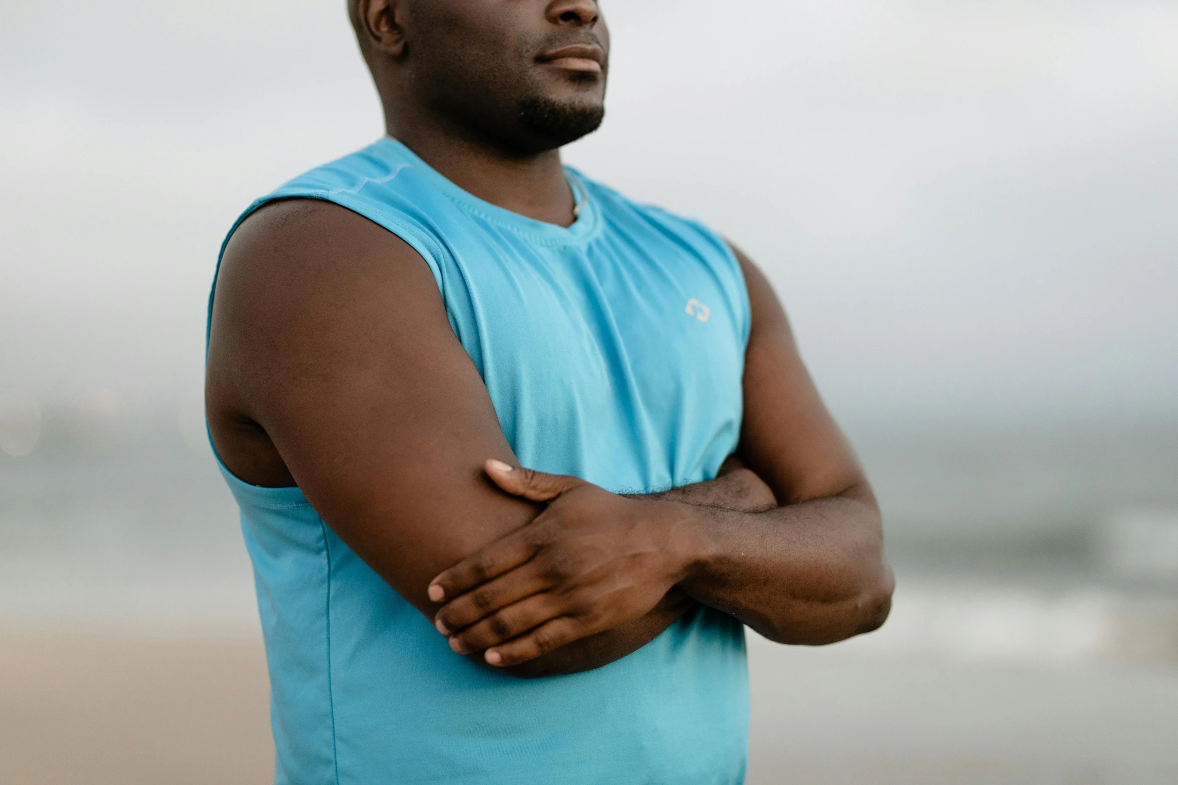 a man standing on a beach with his arms crossed, a portrait, pexels, blue tank top, ( ( dark skin ) ), working out, pensive
