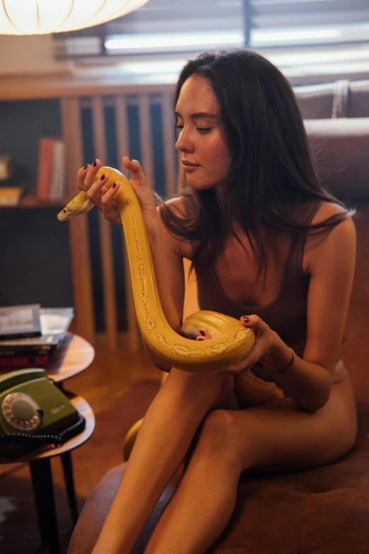 a woman sitting on a couch holding a banana, by Adam Marczyński, trending on pexels, hyperrealism, golden snakes, sexy movie photo, with the head of a snake, brunette