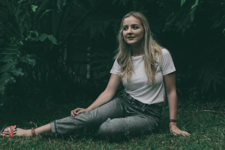 a woman sitting on the grass in front of a bush, a portrait, inspired by Elsa Bleda, pexels contest winner, sophie turner girl, jeans and t shirt, grainy low quality, a blond