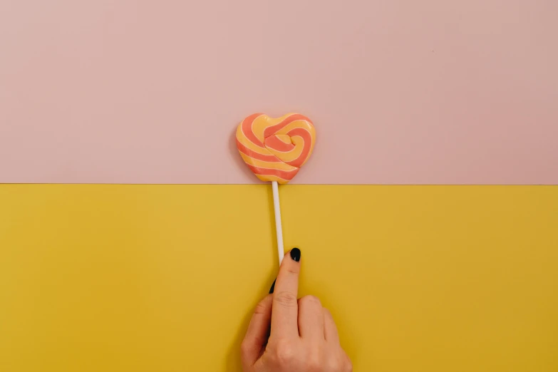 a person holding a lollipop on a yellow and pink background, inspired by Hilma af Klint, trending on pexels, magic heart, 4k polymer clay food photography, on grey background, yellow-orange