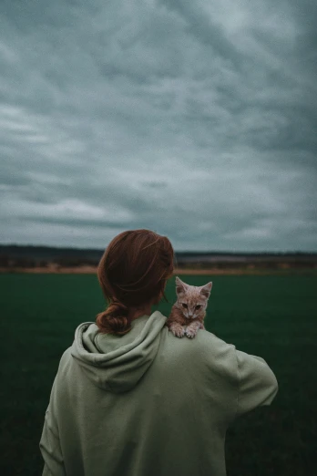 a woman standing in a field with a cat on her shoulder, a picture, trending on pexels, overcast gray skies, ( redhead, a green, square