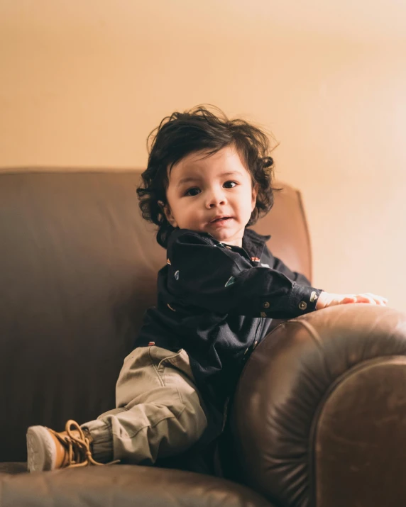 a little boy sitting on top of a brown couch, by Nina Hamnett, pexels contest winner, curls on top, asher duran, wearing jacket, black armchair