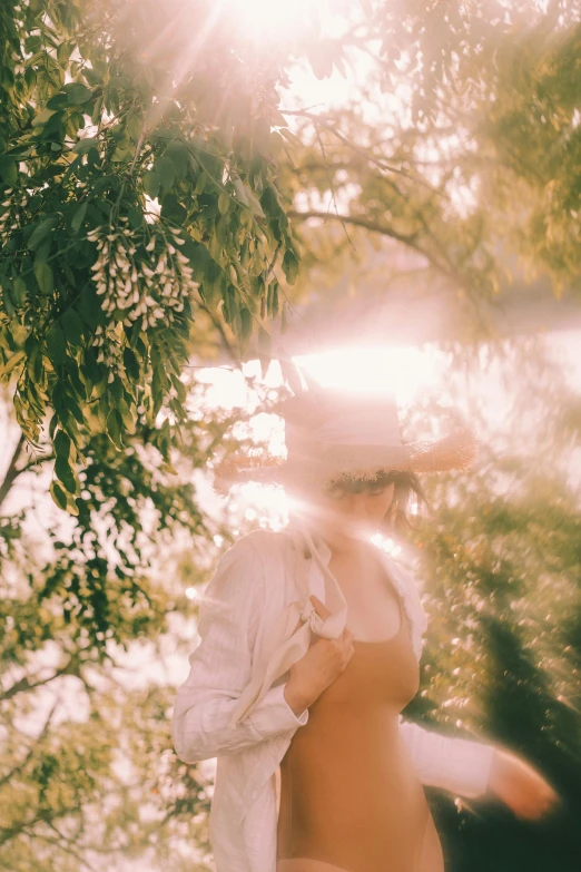 a woman in a bathing suit standing under a tree, unsplash, light and space, light haze, woman with hat, with soft bushes, holy glow