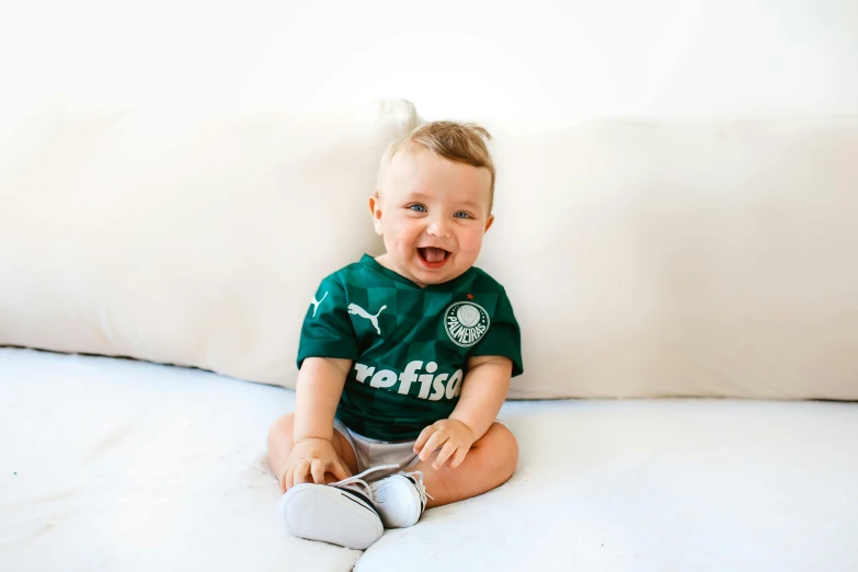 a baby sitting on top of a white couch, inspired by Andrew Robertson, pexels contest winner, happening, celtics, tshirt, smiling spartan, forest green