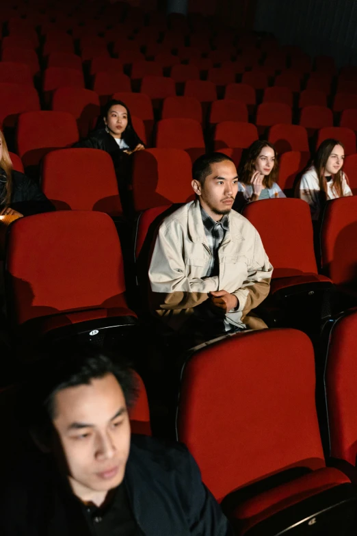 a group of people sitting in rows of red chairs, cinematic movie photograph, [ theatrical ]