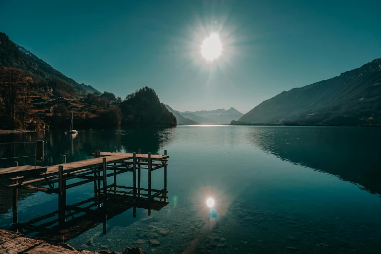 a dock in the middle of a lake with mountains in the background, by Niko Henrichon, pexels contest winner, hurufiyya, with the sun shining on it, sunny day with clear sky, new zeeland, lake blue