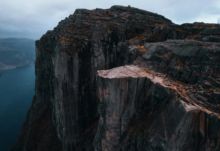 a person standing on the edge of a cliff, by Jesper Knudsen, pexels contest winner, realism, “ aerial view of a mountain, dark grey and orange colours, chiseled formations, scandinavian