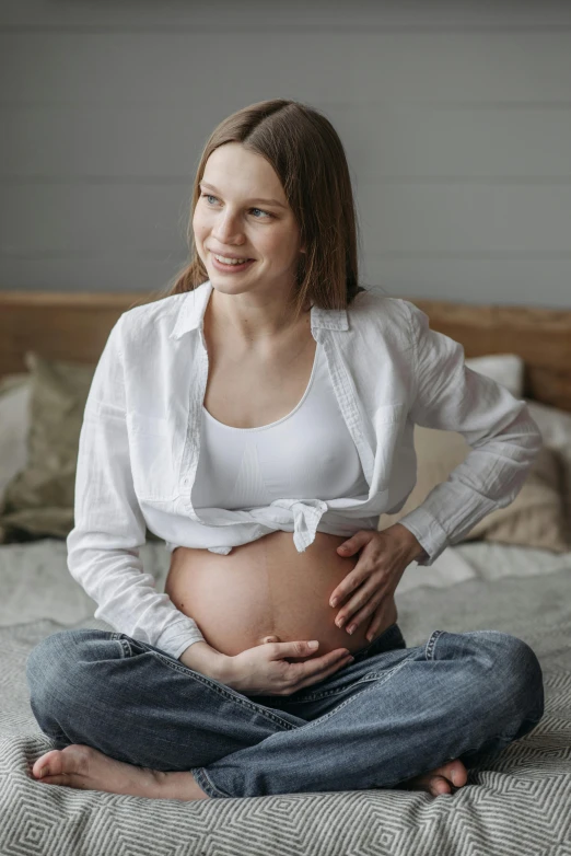 a pregnant woman sitting on top of a bed, wearing a white shirt, profile image, navel, wearing a shirt and a jean