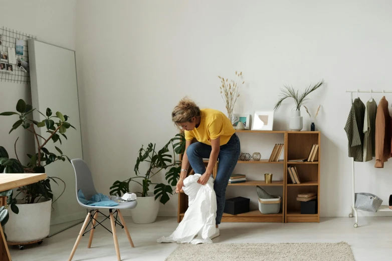 a woman that is standing in a room, inspired by Sarah Lucas, pexels contest winner, happening, picking up a flower, outfit : jeans and white vest, with furniture overturned, lady using yellow dress