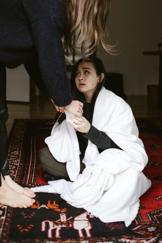 a couple of women sitting on top of a rug, hurting, wearing white robe, recovering from pain, partially cupping her hands