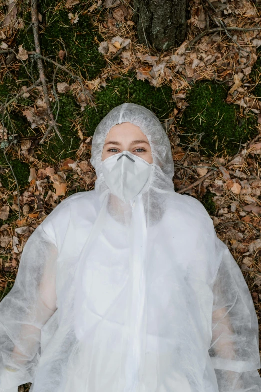 a woman in a white raincoat laying on the ground, inspired by Elsa Bleda, unsplash, environmental art, medical mask, wearing translucent veils, looking the camera, lost in a forest