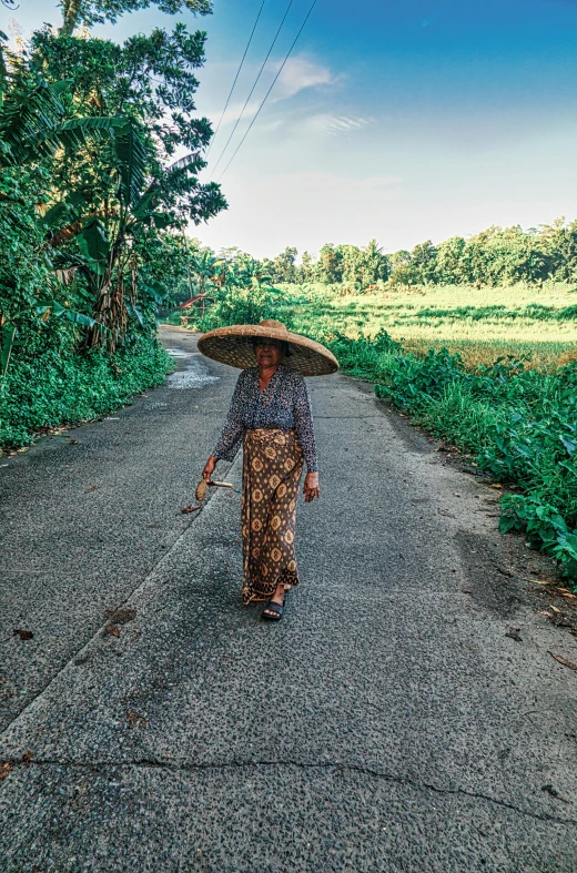a woman walking down a road with a large hat on her head, unsplash, sumatraism, near farm, an oldman, sarong, slightly pixelated