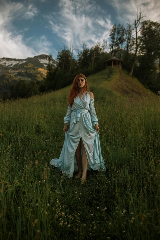 a woman in a blue dress standing in a field, an album cover, by Karl Stauffer-Bern, pexels contest winner, wearing a robe, lauterbrunnen valley, a redheaded young woman, of a full body