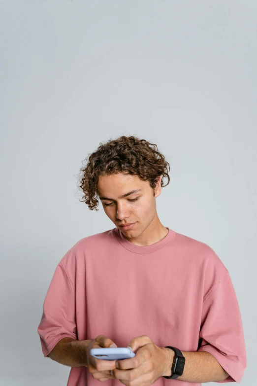 a man in a pink shirt holding a cell phone, a character portrait, trending on pexels, he has short curly brown hair, nonbinary model, shy looking down, wearing sweatshirt