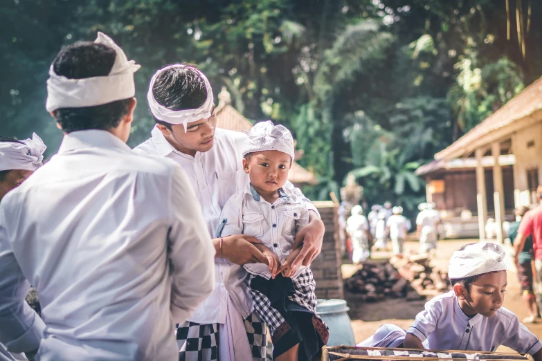 a group of people standing around a table, by Sam Dillemans, pexels contest winner, white sarong, with a kid, ferred - themed robes and hat, arsitektur nusantara