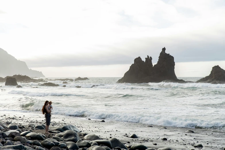 a woman standing on a rocky beach next to the ocean, majestic spires, black sand, family photo, seen from afar