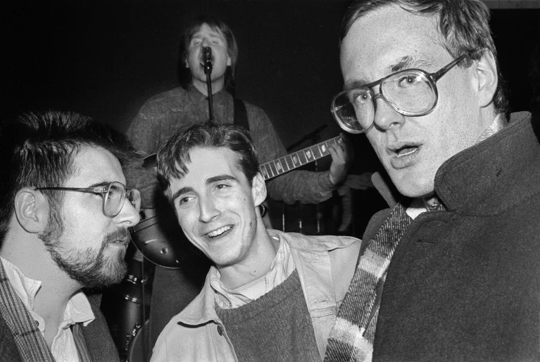 a group of men standing next to each other, an album cover, by Greg Spalenka, art brut, 1 9 8 5 photograph, with nerdy glasses and goatee, wearing a turtleneck and jacket, musicians playing instruments