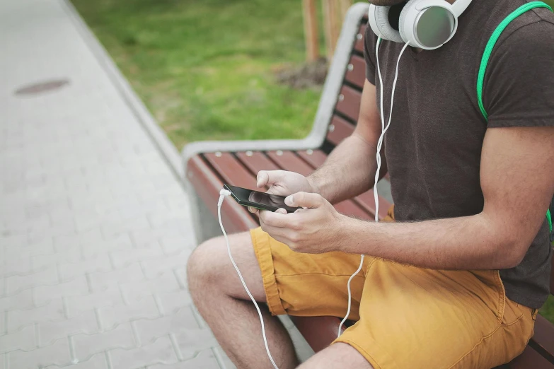 a man sitting on a bench with headphones on, wearing shorts, from reading to playing games, holds a smart phone in one hand, uploaded