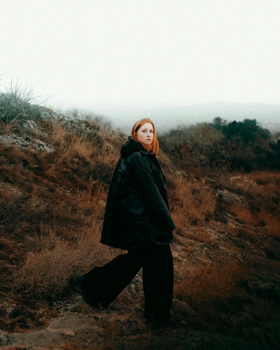 a woman in a black coat standing on a hill, an album cover, unsplash, ginger hair with freckles, low quality photo, full body pictures, view(full body + zoomed out)