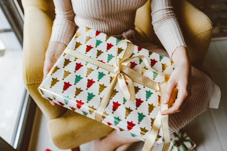 a woman sitting in a chair holding a wrapped gift, by Julia Pishtar, pexels contest winner, arts and crafts movement, emma bridgewater and paperchase, christmas tree, closeup - view, glossy surface