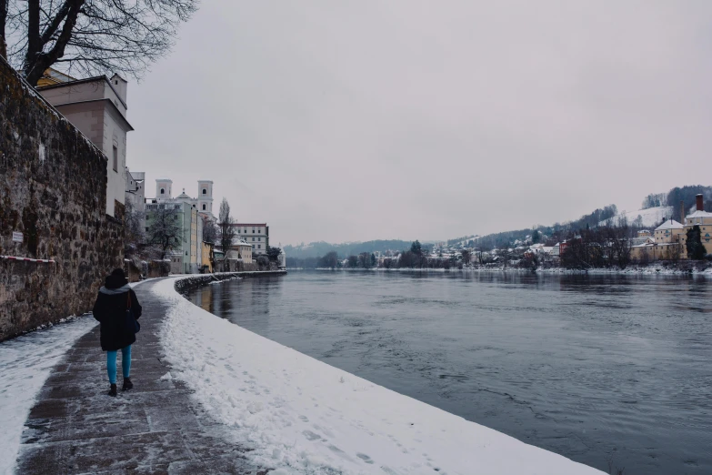 a person walking down a snow covered sidewalk next to a river, inspired by Karl Stauffer-Bern, pexels contest winner, gray skies, hills in the background, 5k, college