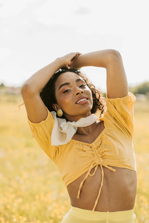 a woman standing in a field with her arms behind her head, trending on pexels, renaissance, wearing yellow croptop, natural complexion, curated collections, flowing golden scarf