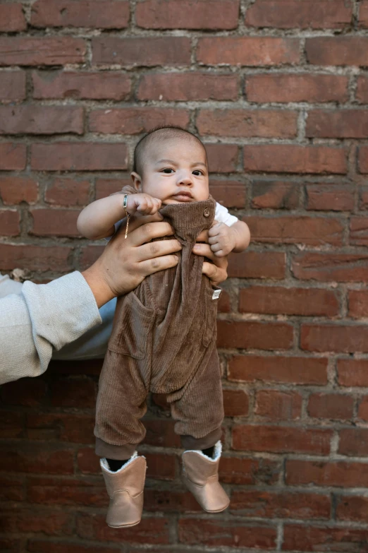 a woman holding a baby in front of a brick wall, by Nina Hamnett, trending on reddit, brown pants, full costume, overalls, brown