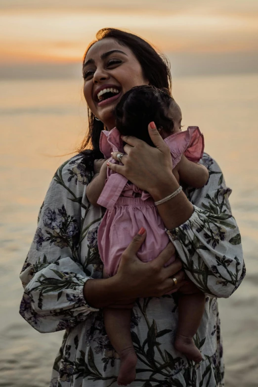 a woman holding a baby next to a body of water, mutahar laughing, pink golden hour, slide show, sri lanka