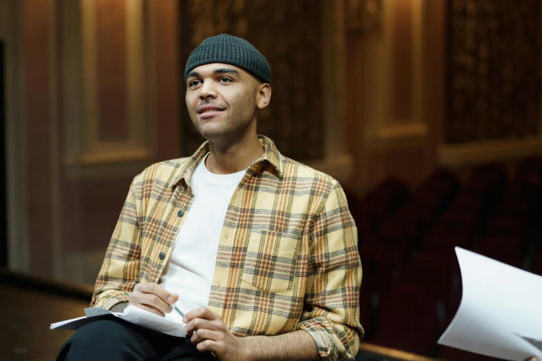 a man sitting in a chair holding a piece of paper, wearing teal beanie, actors, riyahd cassiem, professional image