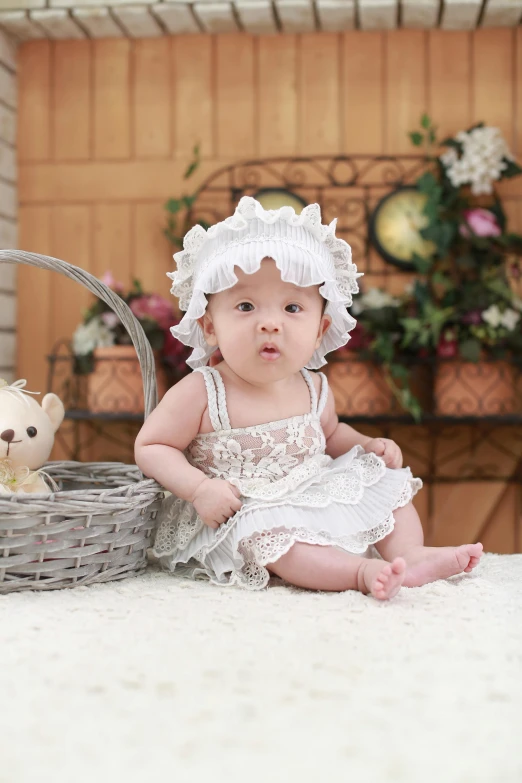 a baby sitting in front of a basket with a teddy bear, a picture, inspired by Margaret Brundage, pixabay contest winner, happening, dressed in a beautiful white, lacey accessories, mai anh tran, with hat