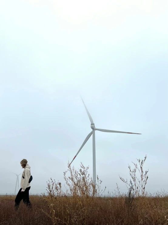 a man walking through a field next to a wind turbine, by Carey Morris, pexels contest winner, under a gray foggy sky, a woman walking, background image, plain background