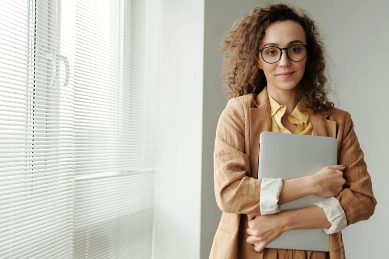 a woman standing in front of a window holding a laptop, trending on pexels, renaissance, nerdy appearance, woman holding another woman, school curriculum expert, in an office