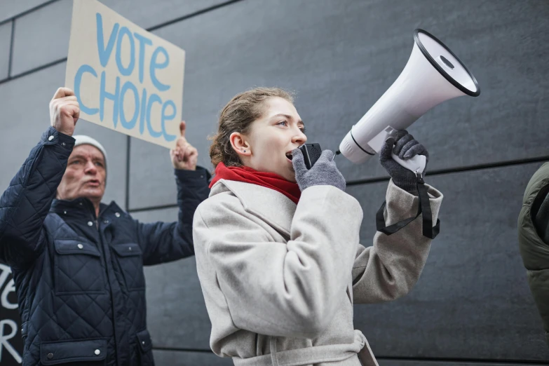 a woman holding a megaphone and a sign, a photo, by Matija Jama, shutterstock, court politics, carice van houten, asking for change, no - text no - logo